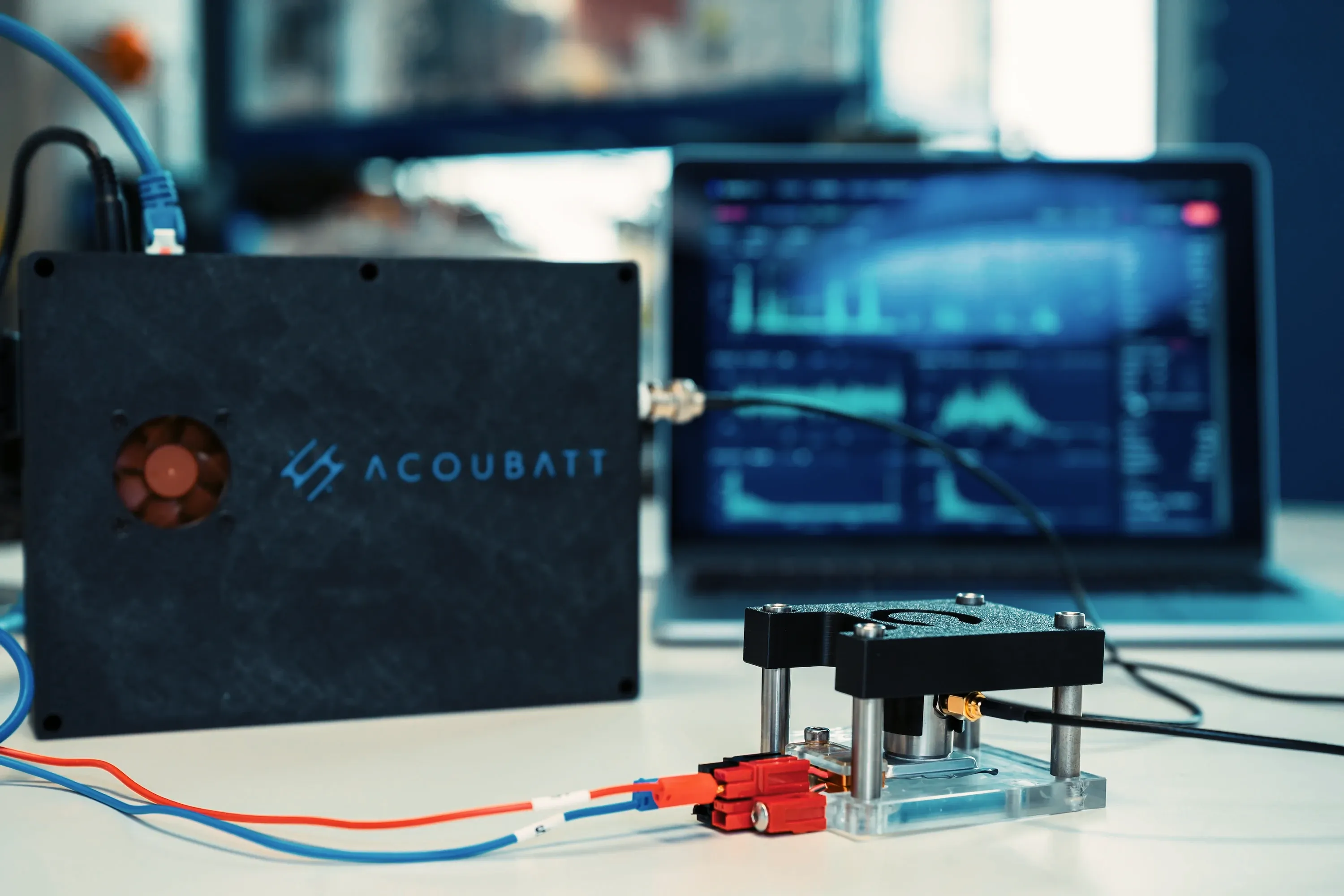 An AcouBatt acquisition unit on a laboratory bench, its sensor lead running to a small cell fixture, with acoustic-emission data on a laptop screen behind.