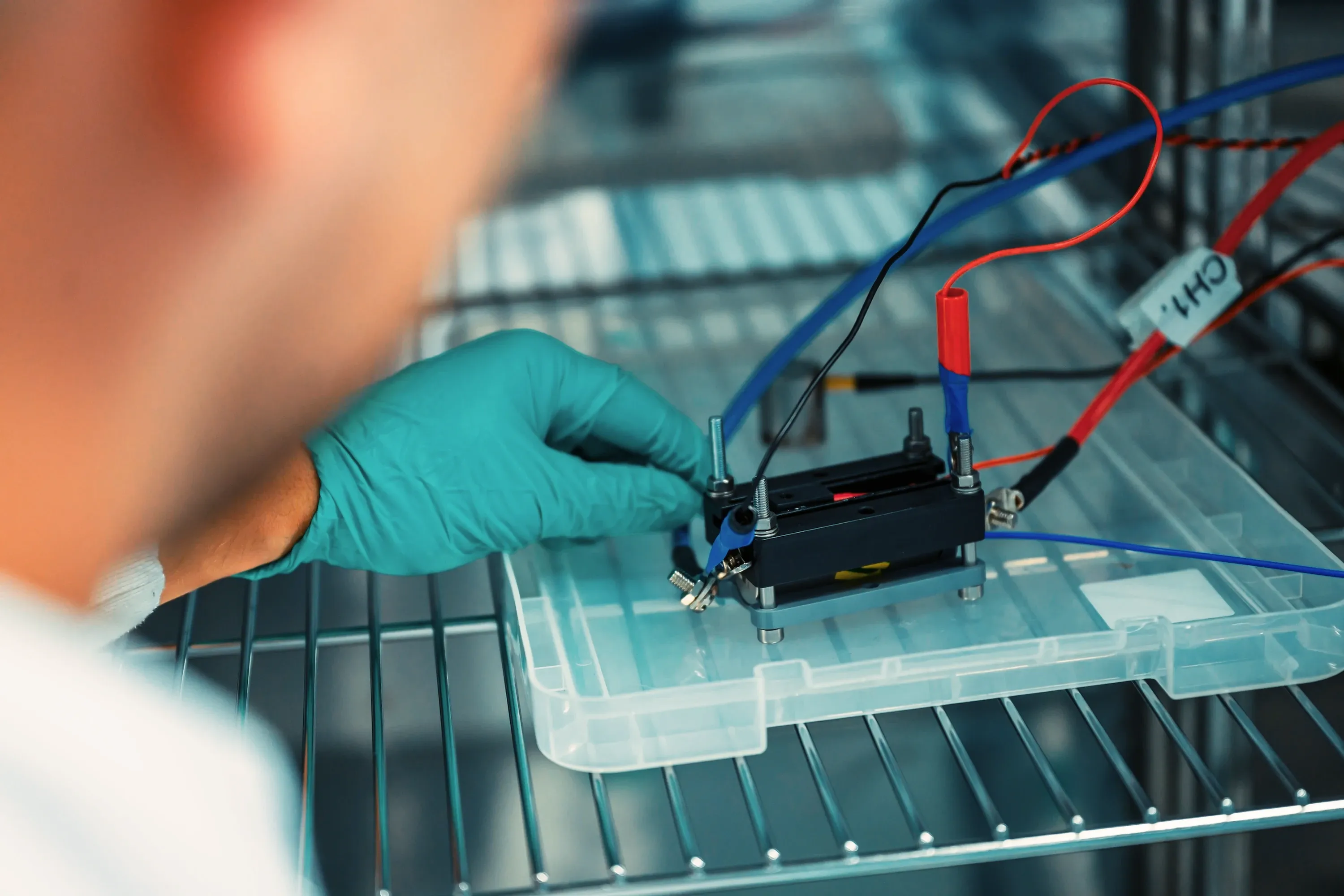 A gloved hand adjusting a cell test fixture on a rack shelf, with its cycler leads attached.