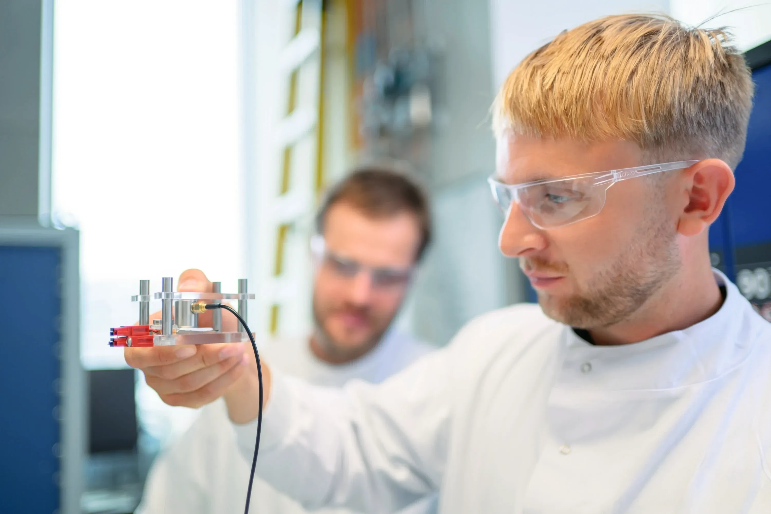 Arthur Fordham holding a small cell fixture with a passive acoustic sensor and its cable, Lukas Noll behind him in the laboratory.