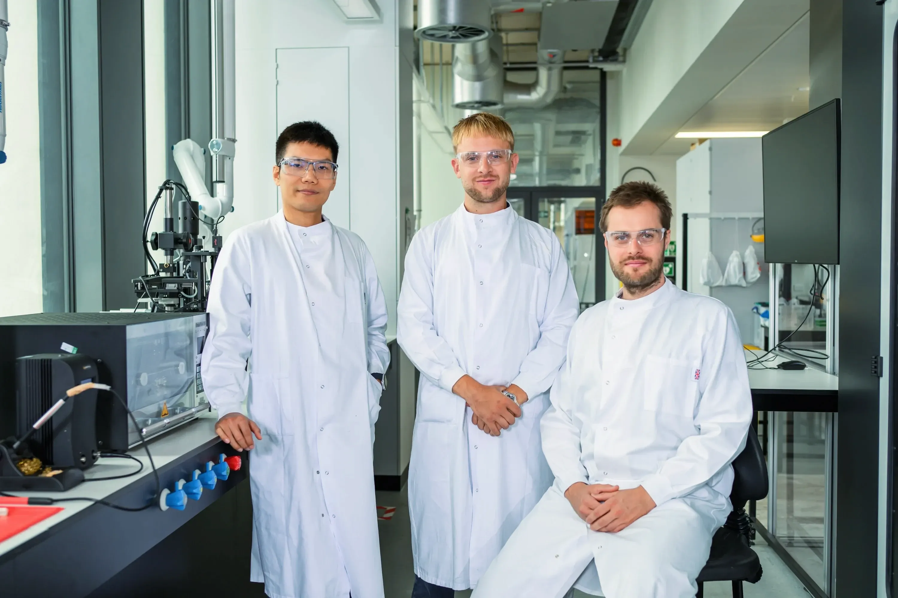 Chris Xu, Arthur Fordham and Lukas Noll in lab coats and safety glasses beside a test bench in the laboratory.
