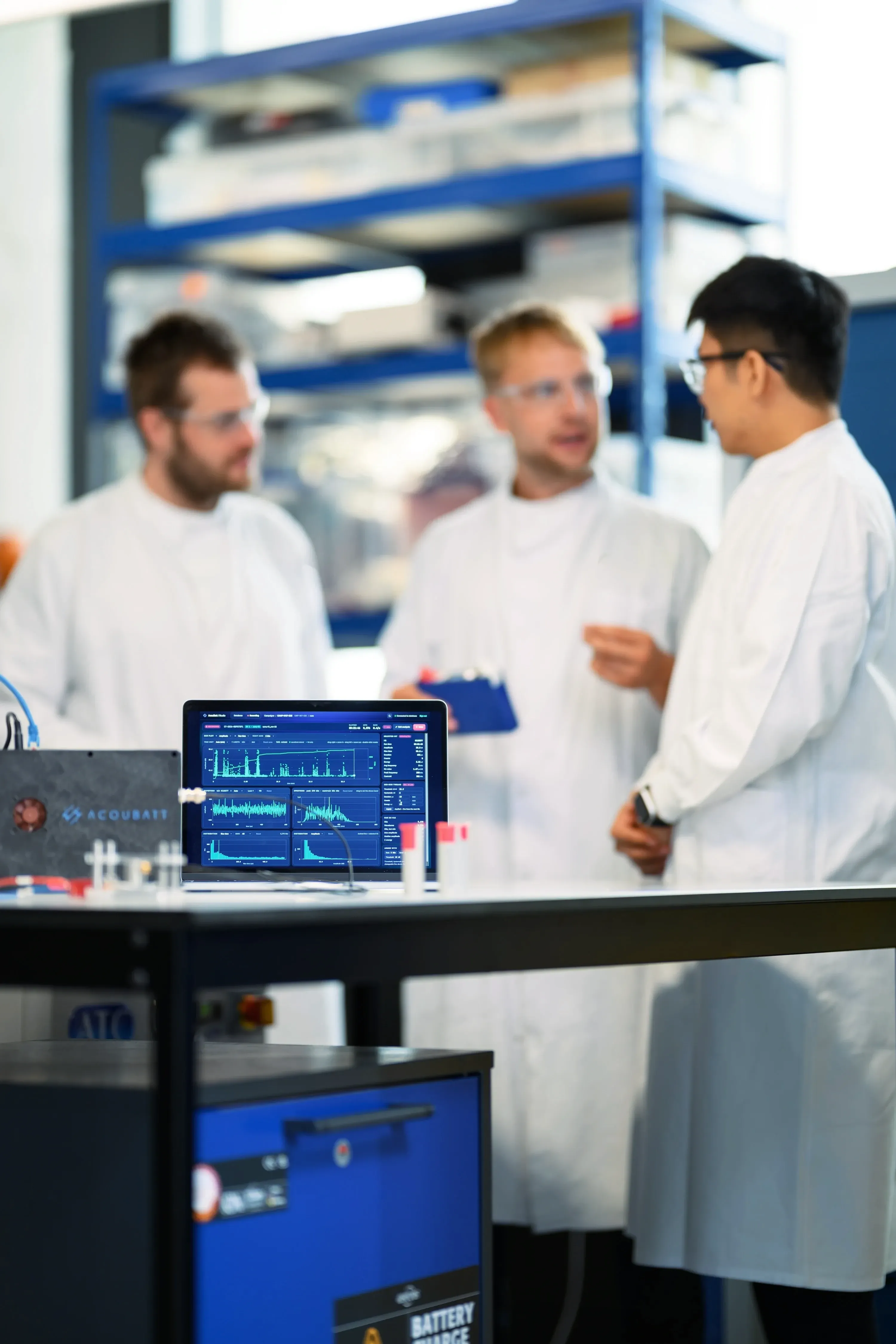 Lukas Noll, Arthur Fordham and Chris Xu in discussion behind a laptop showing AcouBatt's analysis software, with an acquisition unit on the bench in front.