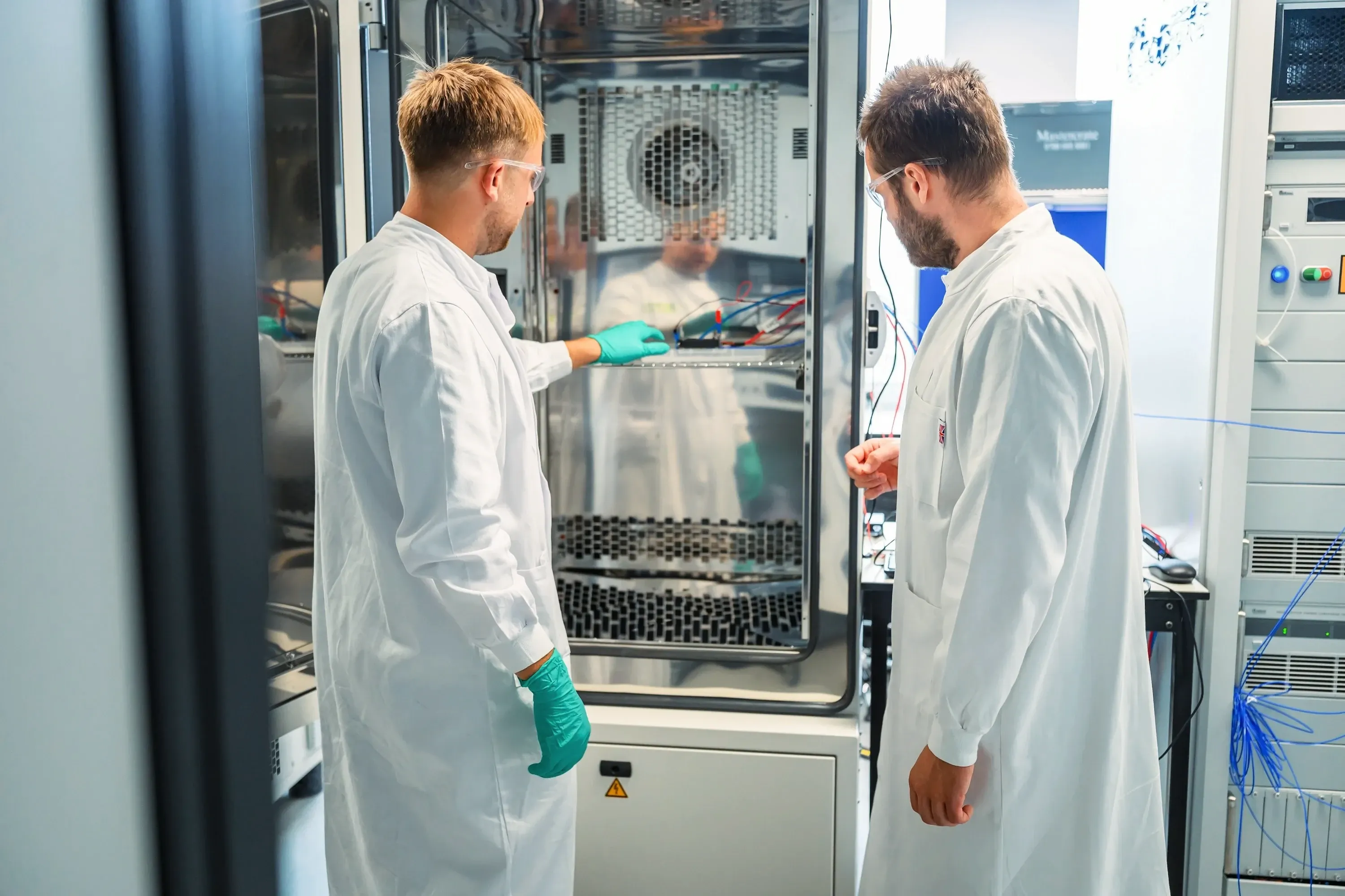 Arthur Fordham and Lukas Noll at an open environmental chamber, checking cells wired to a cycler on its shelf.